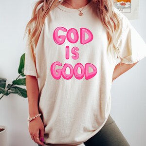Pu&ograve; includere: T-shirt color crema con la scritta "GOD IS GOOD" in lettere rosa a bolle. La maglietta &egrave; indossata con pantaloncini verde scuro. Sono visibili anche un braccialetto e una collana.