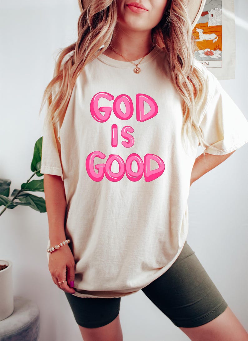 Pu&ograve; includere: T-shirt color crema con la scritta "GOD IS GOOD" in lettere rosa a bolle. La maglietta &egrave; indossata con pantaloncini verde scuro. Sono visibili anche un braccialetto e una collana.