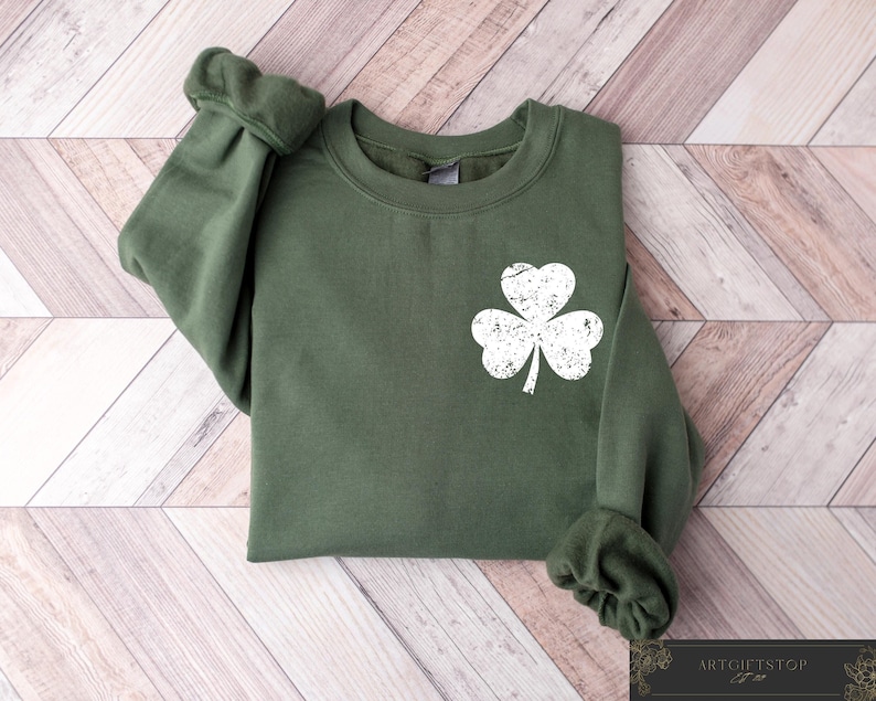 May include: A green sweatshirt with a white, distressed shamrock graphic. The long-sleeved top is made of a soft material and is laid flat on a wooden surface. The cuffs and neckline are ribbed.