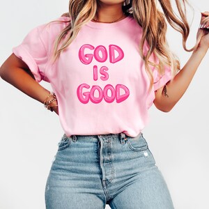 Pu&ograve; includere: Una maglietta rosa chiaro con la scritta "GOD IS GOOD" in lettere a bolle. La maglietta &egrave; abbinata a jeans blu. L'immagine mostra una persona che indossa la maglietta.