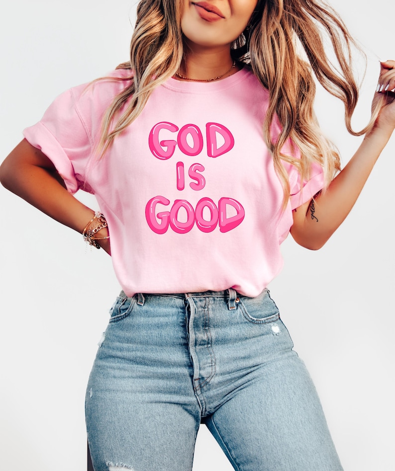 Pu&ograve; includere: Una maglietta rosa chiaro con la scritta "GOD IS GOOD" in lettere a bolle. La maglietta &egrave; abbinata a jeans blu. L'immagine mostra una persona che indossa la maglietta.