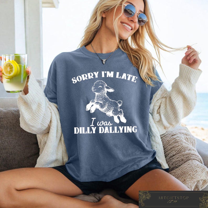 May include: A blue t-shirt with the text "SORRY I'M LATE I WAS DILLY DALLYING" and a white graphic of a jumping lamb. The shirt is worn by a person wearing sunglasses and a cream-colored cardigan.