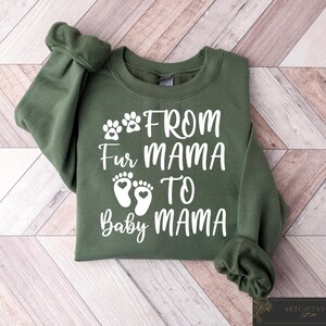 May include: An olive green sweatshirt with the text "From Fur Mama To Baby Mama" in white. The design includes paw prints and baby footprints. The sweatshirt is folded on a light brown and white wooden surface.