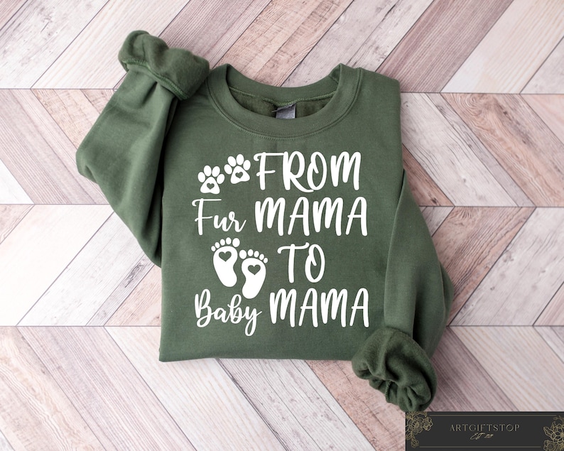 May include: An olive green sweatshirt with the text "From Fur Mama To Baby Mama" in white. The design includes paw prints and baby footprints. The sweatshirt is folded on a light brown and white wooden surface.
