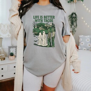 以下が含まれることがあります： 「LIFE IS BETTER WITH TACOS」と書かれた緑色のグラフィックが特徴のグレーのTシャツ。グラフィックは、タコトラック、幽霊、ゾンビが描かれています。このシャツはカジュアルな半袖スタイルです。