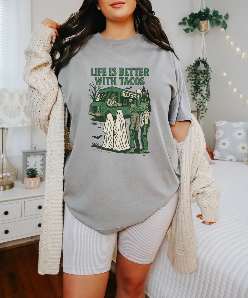 以下が含まれることがあります： 「LIFE IS BETTER WITH TACOS」と書かれた緑色のグラフィックが特徴のグレーのTシャツ。グラフィックは、タコトラック、幽霊、ゾンビが描かれています。このシャツはカジュアルな半袖スタイルです。