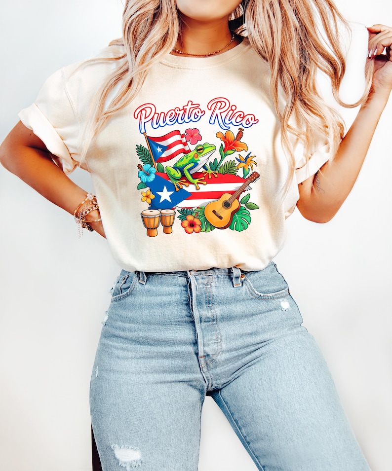 May include: Cream-colored t-shirt featuring a colorful graphic of the Puerto Rican flag, a frog, flowers, a guitar, and the text "Puerto Rico". The design includes bongo drums and tropical foliage. The shirt is paired with light blue jeans.