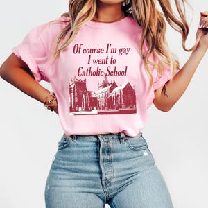 May include: Pink t-shirt with a red graphic of a church and the text "Of course I'm gay I went to Catholic School."