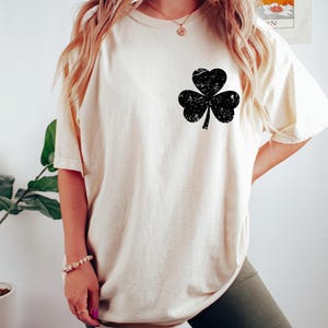 May include: Cream-colored t-shirt featuring a distressed black shamrock design on the front. The shirt has short sleeves and a relaxed fit. A St. Patrick's Day themed apparel item.