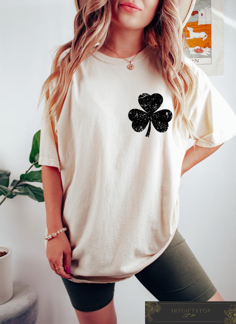 May include: Cream-colored t-shirt featuring a distressed black shamrock design on the front. The shirt has short sleeves and a relaxed fit. A St. Patrick's Day themed apparel item.