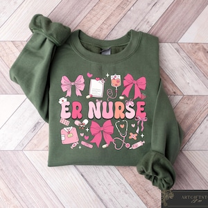 May include: Green sweatshirt with a pink and white graphic design that says "ER Nurse". The graphic includes hearts, bandages, a stethoscope, and other medical symbols.