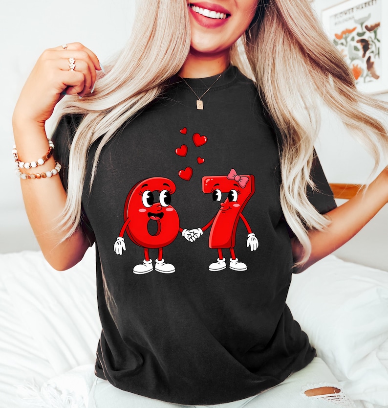 May include: Black t-shirt featuring a cartoon design of the numbers 6 and 7 holding hands, with hearts floating above them. The numbers are red with white accents, and the number 7 has a pink bow. The design is centered on the shirt.