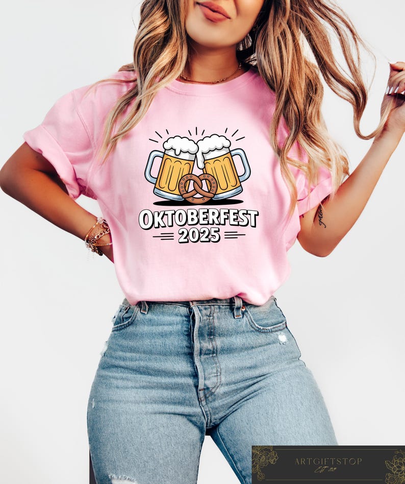 May include: Pink t-shirt featuring a graphic of two beer steins with foamy tops and a pretzel, with the text "OKTOBERFEST 2025". The shirt is a light pink color.