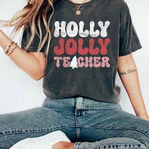 Op de afbeelding: Donkergrijs t-shirt met de tekst "HOLLY JOLLY TEACHER" in wit, rood en roze, met een kerstboomafbeelding. Het shirt wordt gedragen met een blauwe jeans en witte sneakers. Een gouden ketting en armband zijn ook zichtbaar.