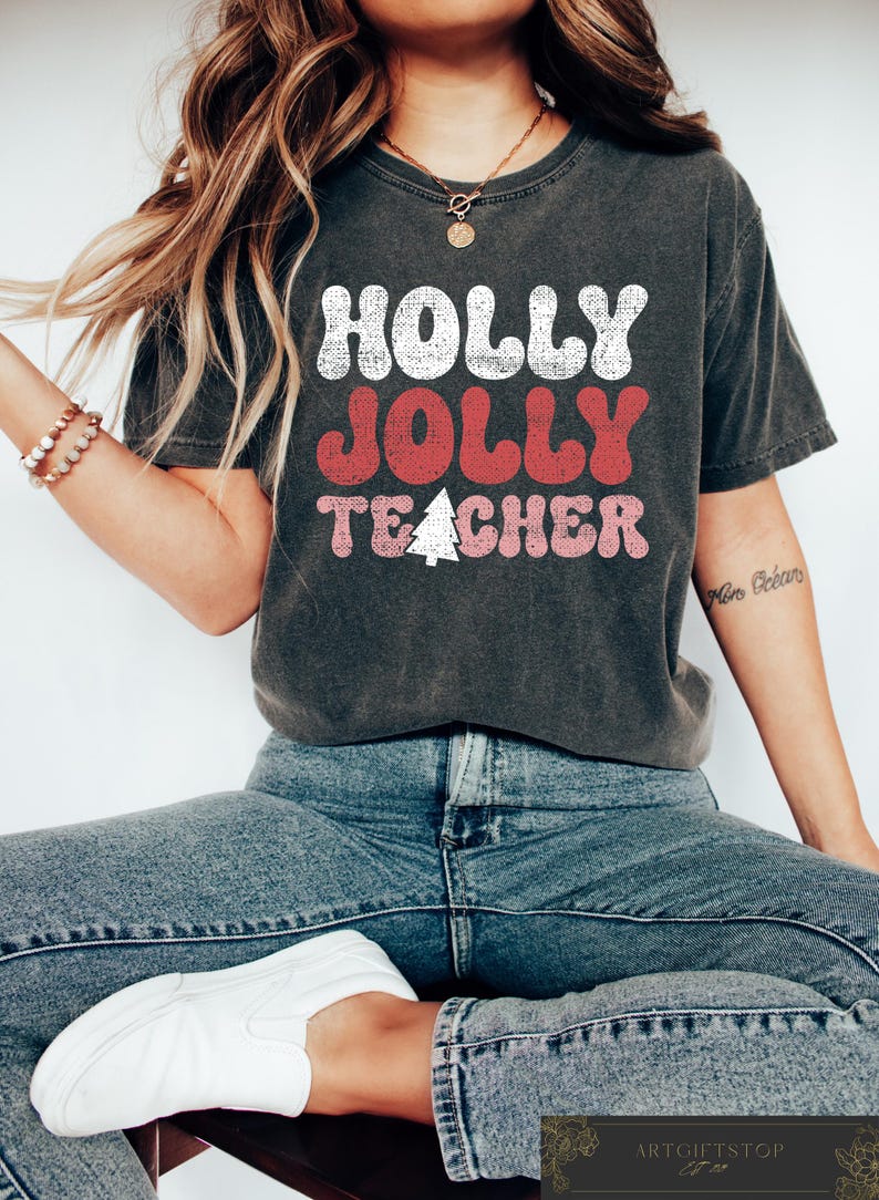 Op de afbeelding: Donkergrijs t-shirt met de tekst "HOLLY JOLLY TEACHER" in wit, rood en roze, met een kerstboomafbeelding. Het shirt wordt gedragen met een blauwe jeans en witte sneakers. Een gouden ketting en armband zijn ook zichtbaar.