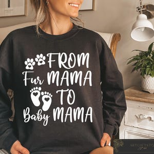 May include: Black sweatshirt with white text that reads "FROM Fur MAMA TO Baby MAMA". The design includes paw prints and baby footprints with hearts. The sweatshirt is a casual style.