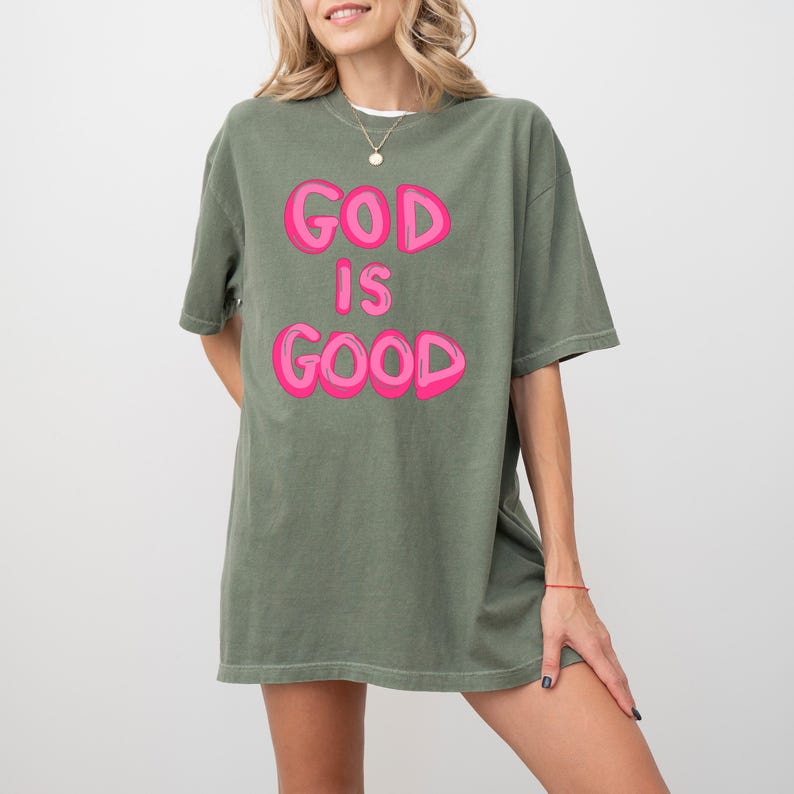 Pu&ograve; includere: Una maglietta verde oliva con la scritta "GOD IS GOOD" in lettere rosa acceso. La maglietta ha maniche corte e una vestibilit&agrave; ampia. La modella indossa una collana d'oro e un braccialetto rosso.
