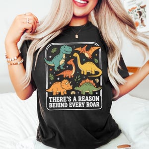 May include: Black t-shirt featuring a colorful dinosaur graphic. The design includes various dinosaurs in shades of green, orange, and yellow, with the text "THERE'S A REASON BEHIND EVERY ROAR" in a white rectangular frame.