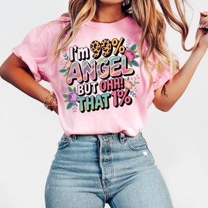 Op de afbeelding: Roze t-shirt met de tekst "I'm 99% ANGEL BUT OHH! THAT 1%" in een kleurrijk, bloemenontwerp. Het shirt heeft korte mouwen en is gemaakt van een zacht materiaal. Het ontwerp bevat een luipaardprint en pastelkleuren.