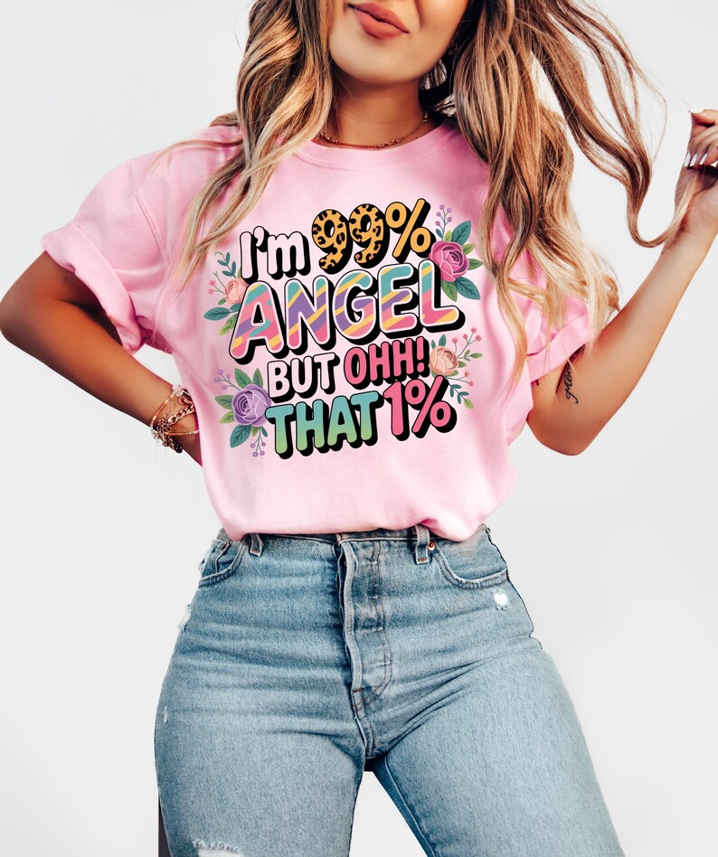 Op de afbeelding: Roze t-shirt met de tekst "I'm 99% ANGEL BUT OHH! THAT 1%" in een kleurrijk, bloemenontwerp. Het shirt heeft korte mouwen en is gemaakt van een zacht materiaal. Het ontwerp bevat een luipaardprint en pastelkleuren.