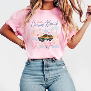 May include: Pink t-shirt with the words "Cousins Beach" and a graphic of a yellow truck carrying a surfboard, surrounded by beach-themed illustrations. The text "We will always have summer" is also included.