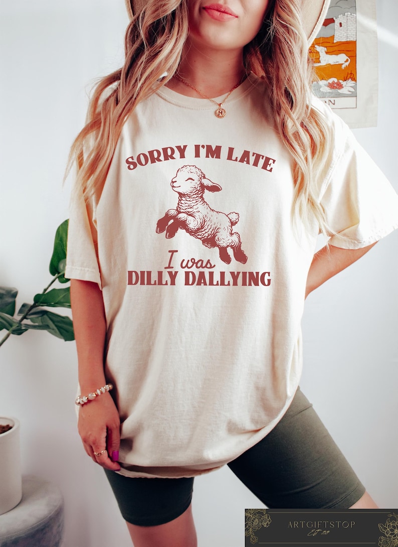 May include: Cream-colored t-shirt with the text "SORRY I'M LATE I was DILLY DALLYING" in maroon. A cartoon lamb is jumping in the center. The shirt is a relaxed fit.