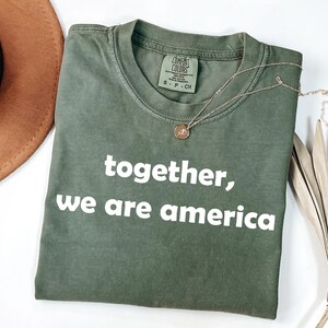 Puede incluir: Una camiseta verde oliva con el texto "together, we are america" en blanco. Un collar dorado con un colgante descansa sobre la camiseta. Un sombrero marrón y una hoja de palma seca están en el fondo.