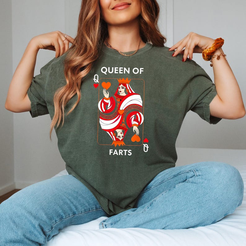 May include: Olive green t-shirt featuring a playing card design with the text "QUEEN OF FARTS". The card depicts a queen with a crown, red and white details, and heart symbols. The shirt is worn by a person in blue jeans.