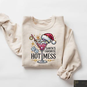 May include: A beige sweatshirt with a festive design featuring a martini glass with red liquid and ice cubes, a Santa hat, and the text "Santa's Favorite Hot Mess".