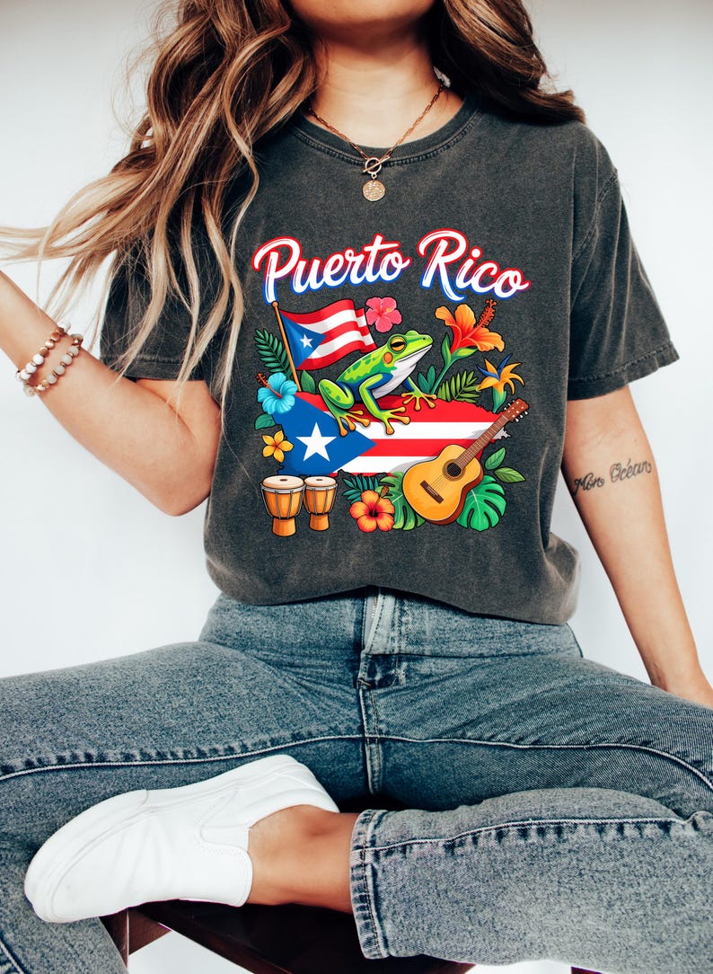 May include: Dark gray t-shirt with the text "Puerto Rico" in red and blue script. The graphic includes a frog, flag, flowers, guitar, and drums, all in vibrant colors. The shirt is a comfortable, casual style.