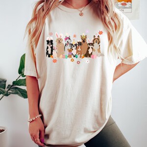 May include: A white t-shirt with a graphic of dogs wearing bunny ears and holding Easter eggs. The dogs are all different breeds and colors. The graphic is in a cartoon style and is surrounded by flowers.