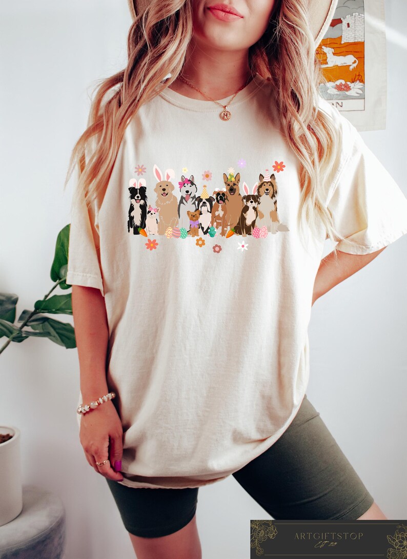 May include: A white t-shirt with a graphic of dogs wearing bunny ears and holding Easter eggs. The dogs are all different breeds and colors. The graphic is in a cartoon style and is surrounded by flowers.