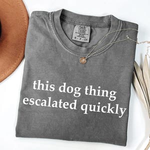 May include: A charcoal grey short-sleeve t-shirt with a crew neckline and the white text "this dog thing escalated quickly" printed on the front. The shirt features a Comfort Colors brand label at the inner neck and is made of cotton fabric.