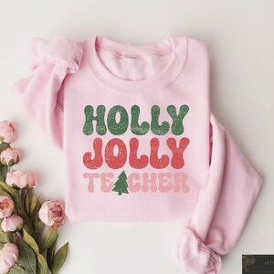 Op de afbeelding: Een lichtroze sweatshirt met de tekst "HOLLY JOLLY TEACHER" in groen, rood en roze. Een kleine groene kerstboomafbeelding staat onder het woord "TEACHER". Het sweatshirt is op een wit oppervlak met roze bloemen gevouwen.