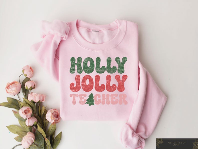 Op de afbeelding: Een lichtroze sweatshirt met de tekst "HOLLY JOLLY TEACHER" in groen, rood en roze. Een kleine groene kerstboomafbeelding staat onder het woord "TEACHER". Het sweatshirt is op een wit oppervlak met roze bloemen gevouwen.