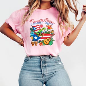 May include: Pink t-shirt with a colorful graphic featuring the words "Puerto Rico" above a frog, flag, guitar, and flowers. The design incorporates the Puerto Rican flag and musical instruments.