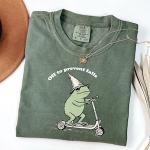 May include: Olive green t-shirt with the phrase "Off to prevent falls" above a cartoon frog wearing a wizard hat and riding a scooter. The shirt has a crew neck and short sleeves. A gold necklace is draped across the shirt.