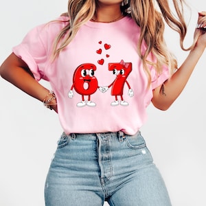 May include: Pink t-shirt featuring a cartoon design of the numbers six and seven holding hands, with hearts floating above them. The numbers have faces, gloves, and shoes. A bow adorns the number seven.