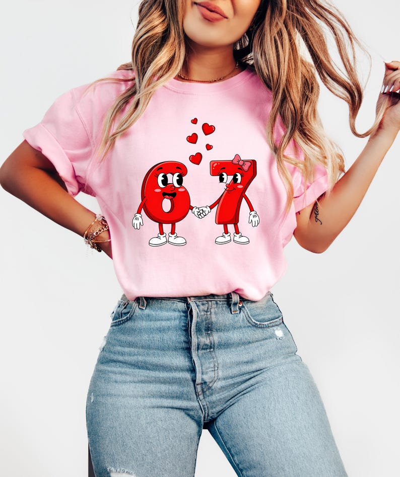 May include: Pink t-shirt featuring a cartoon design of the numbers six and seven holding hands, with hearts floating above them. The numbers have faces, gloves, and shoes. A bow adorns the number seven.