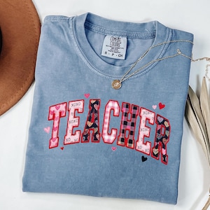 May include: A blue Comfort Colors t-shirt with the word "TEACHER" embroidered in a collegiate style. The letters feature heart and plaid patterns, with "XOXO" and heart accents. A gold necklace and a brown hat are also visible.