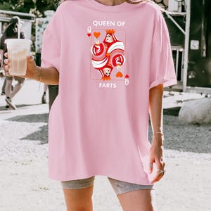 May include: A light pink t-shirt featuring a playing card design with a queen holding a heart. The text on the shirt reads "QUEEN OF FARTS". The shirt is a loose fit.