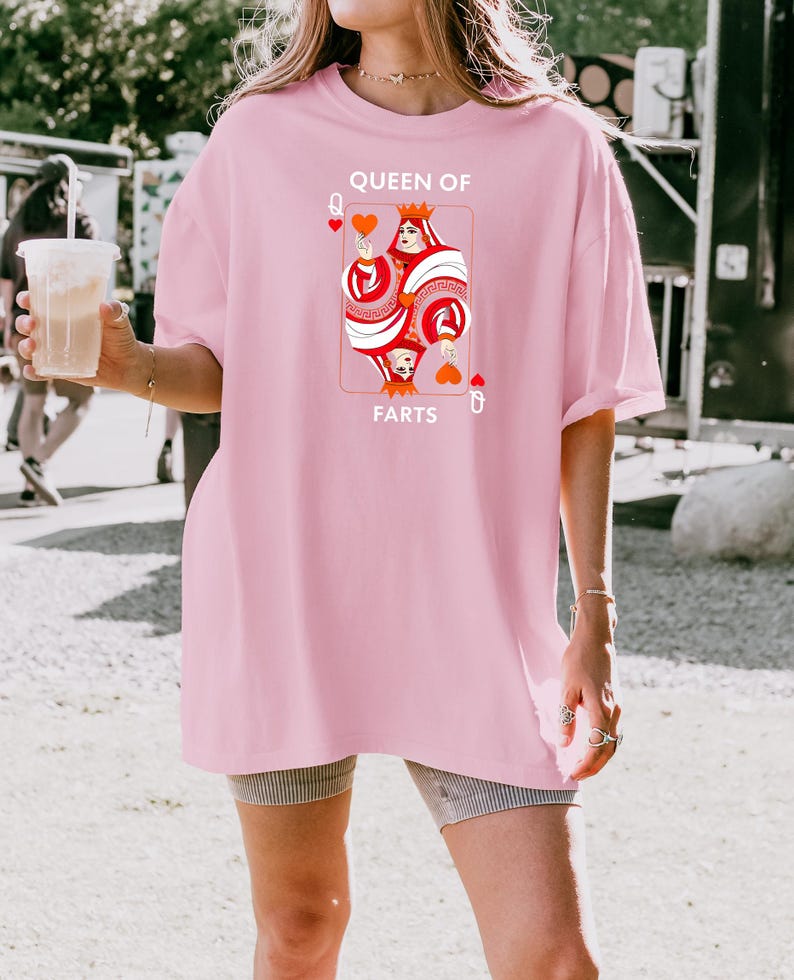 May include: A light pink t-shirt featuring a playing card design with a queen holding a heart. The text on the shirt reads "QUEEN OF FARTS". The shirt is a loose fit.