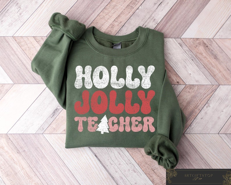 Op de afbeelding: Een groene sweatshirt met de tekst "HOLLY JOLLY TEACHER" in wit, rood en roze, met een witte kerstboomafbeelding. De sweatshirt wordt gepresenteerd op een houten oppervlak.