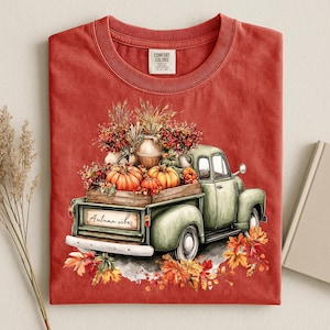 Könnte beinhalten: Ein ziegelrotes T-Shirt mit einer Grafik eines hellgrünen Oldtimer-Pickups, der Kürbisse, Weizen und Herbstlaub auf der Ladefläche trägt. Auf der Heckklappe steht der Text Autumn vibes in Schreibschrift. Das Shirt hat einen Rundhalsausschnitt.