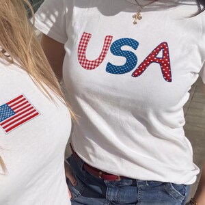 May include: Two white t-shirts with patriotic designs. One has an American flag patch, the other spells "USA" with red and blue patterned letters. Both are worn with denim skirts.