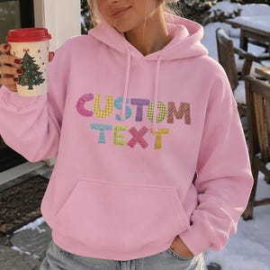 May include: A light pink hooded sweatshirt featuring the words CUSTOM TEXT in multi-colored, patterned, textured block letters across the chest. The hoodie includes a front kangaroo pocket, long sleeves, and a drawstring hood.