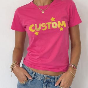 May include: A bright pink short-sleeved t-shirt featuring the word CUSTOM printed in a yellow checkered pattern across the chest, accented by three small yellow stars positioned above and below the text, paired with a blue denim mini skirt.
