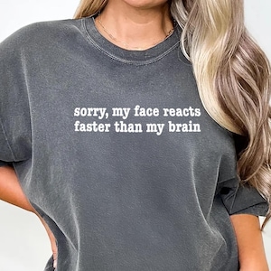 May include: Dark gray t-shirt with the text "sorry, my face reacts faster than my brain" in white. The shirt has a relaxed fit and short sleeves. The shirt is a casual wear item.