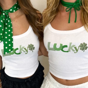 Puede incluir: Dos mujeres con camisetas sin mangas blancas con la palabra "Lucky" en verde y un trébol verde. Una lleva una falda de encaje negra y una bufanda verde con lunares. La otra lleva una falda de encaje blanca y una gargantilla verde.
