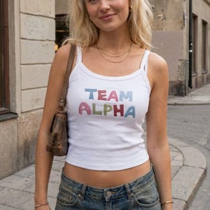 以下が含まれることがあります： 青、ピンク、緑で「TEAM ALPHA」と書かれた白いタンクトップ。細いストラップが付いており、ブルージーンズと合わせています。茶色のショルダーバッグが肩からかけてあります。